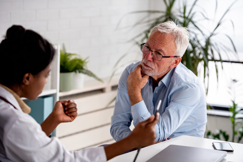 A healthcare professional gestures while explaining a medical document to an older man during a clinic consultation. They sit at a white desk with a laptop and smartphone nearby.