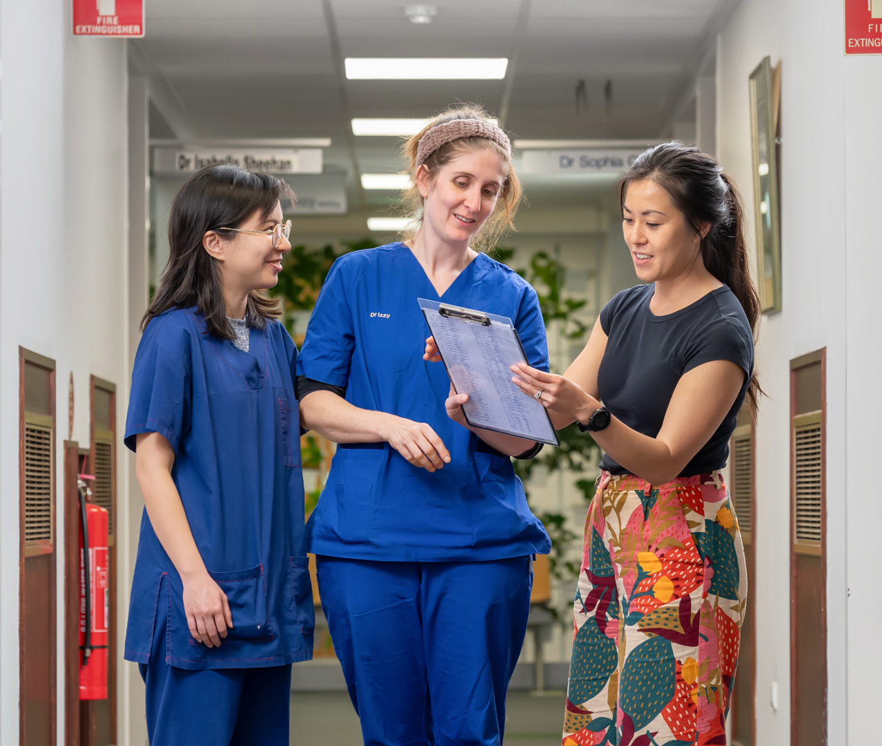 Three women stand in a hallway having a discussion while looking at a clipboard. Two women wear medical scrubs, and one wears a black top with a colourful patterned skirt.
