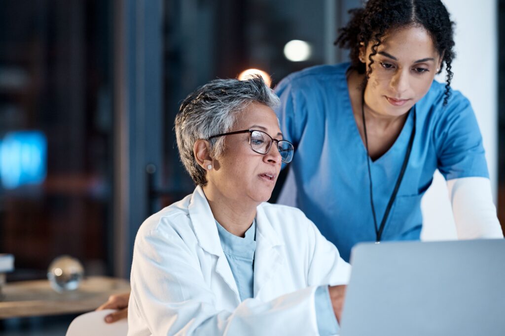 Two female healthcare professionals, one in a white coat and the other in blue scrubs, review information on a laptop together. The setting is a medical office.