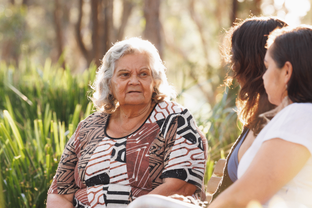 An older Aboriginal woman sits outside in natural bushland surroundings, speaking with two younger women. She wears a patterned dress and appears engaged in the conversation under soft afternoon light.