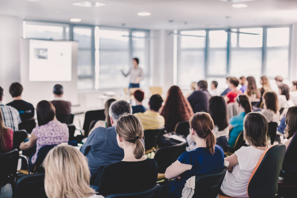 A diverse group of adults sits in rows listening to a presenter at the front of a brightly lit room. A projector screen is visible in the background as the presenter gestures while speaking.