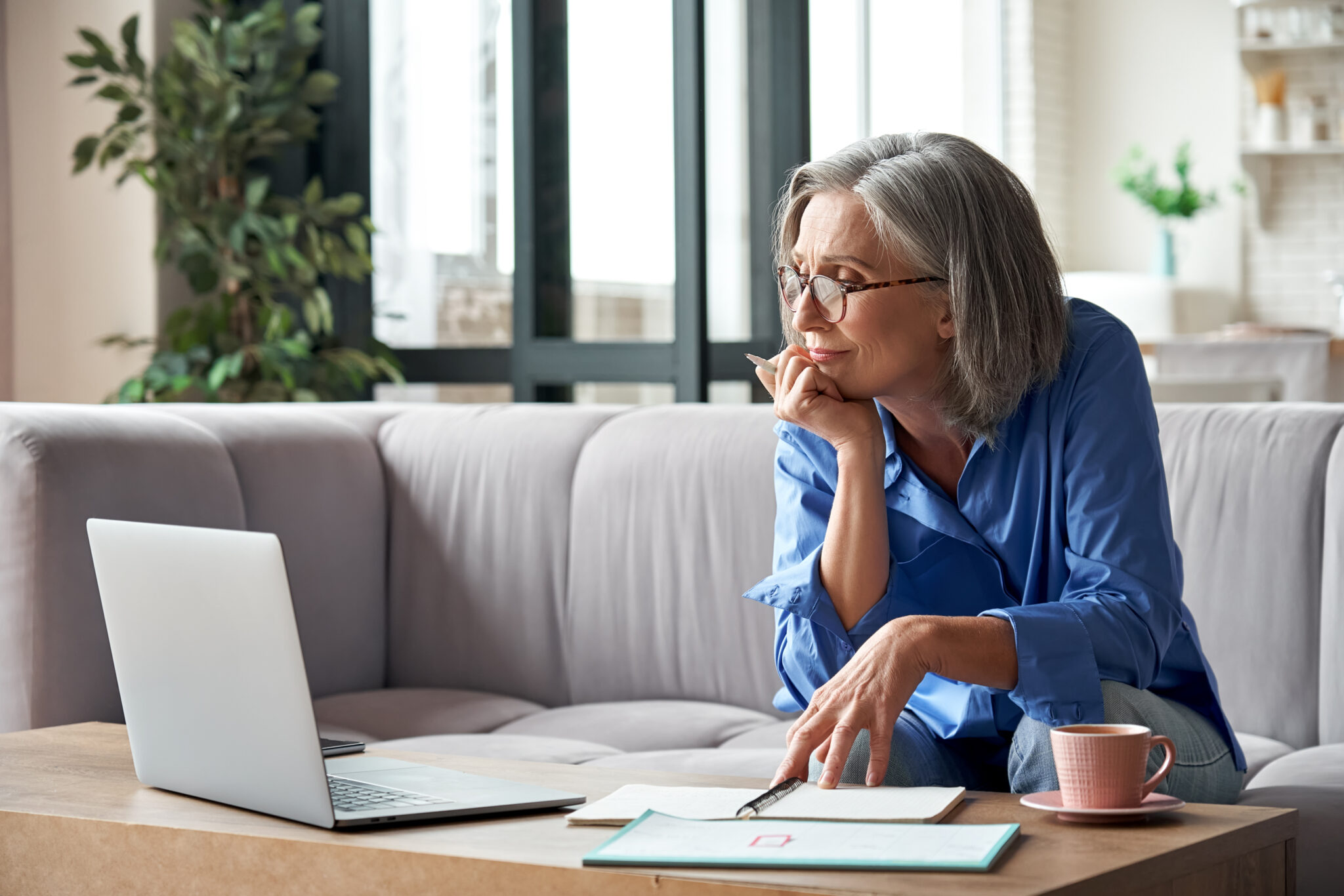 A woman with grey hair and glasses sits on a couch, taking notes while looking at her laptop. A notebook, pen, and coffee cup sit on the table in front of her.