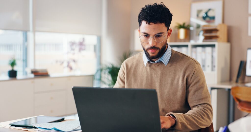 A young man with glasses and a beard works at a laptop in a bright, modern office. He wears a tan jumper and sits at a desk surrounded by documents, a tablet, and office supplies.