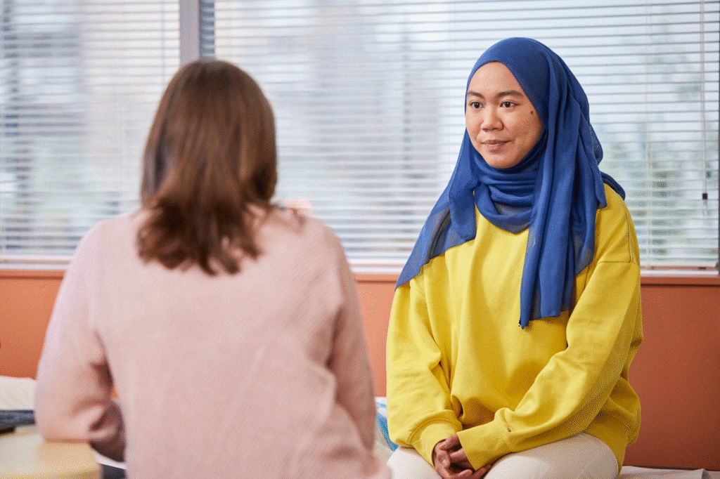 A young woman wearing a blue headscarf and yellow jumper sits across from another woman during a conversation. They are in a room with closed blinds and a calm, neutral tone.